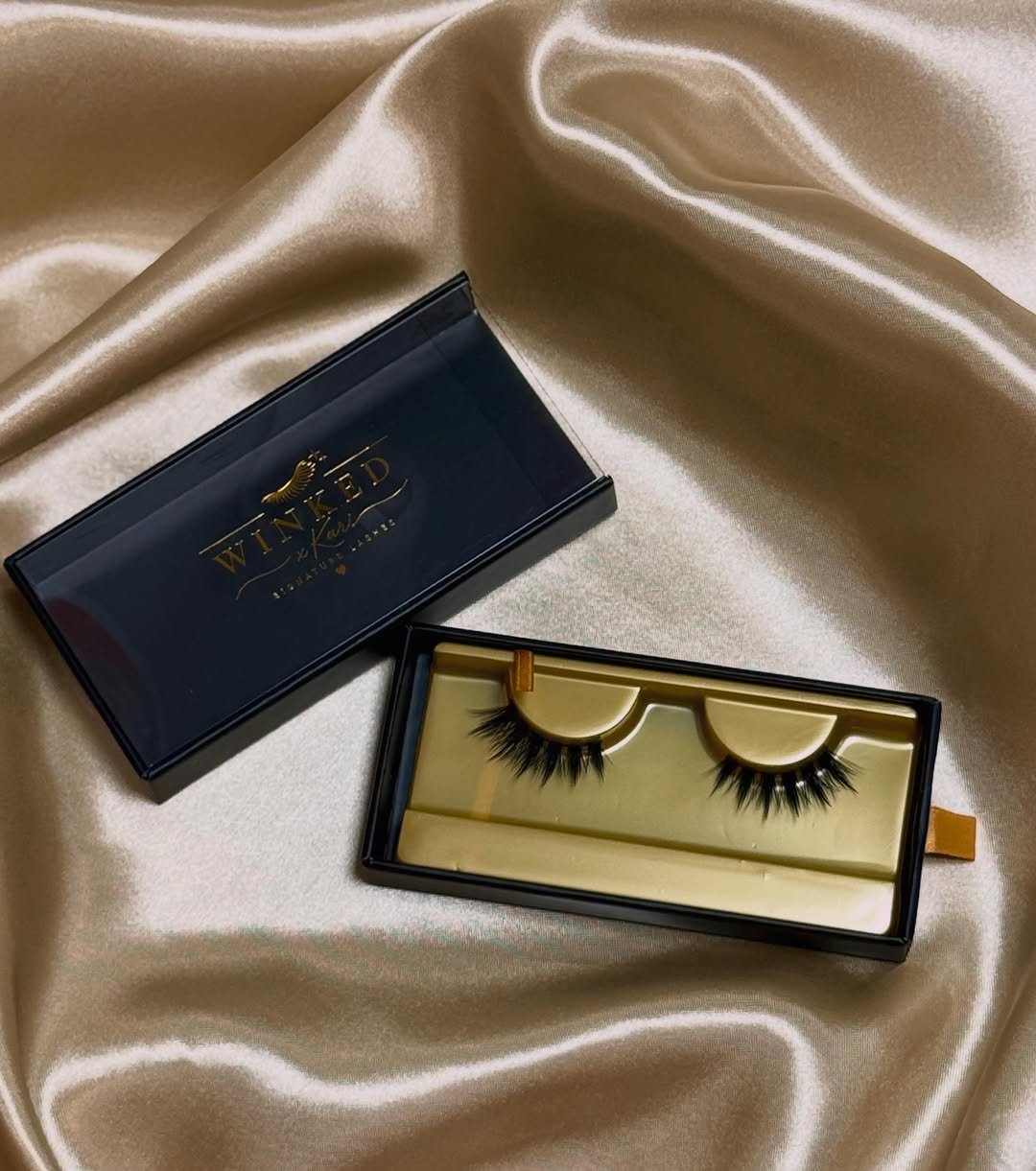 Black false eyelashes in a gold and black Winked x Kari box on draped beige satin